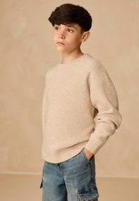 Beżowy sweter z dzianiny o fakturowym wzorze, z okrągłym dekoltem i ściśniętymi mankietami, zestawiony z jasnoniebieskimi spodniami cargo z denimu. Prosty design.