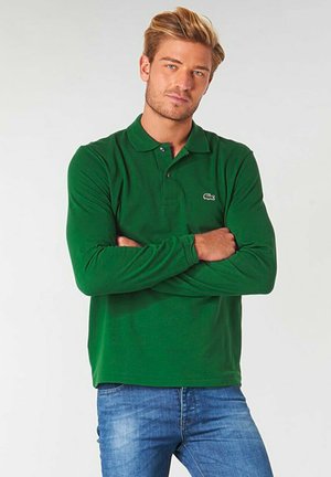 Polo a maniche lunghe verde con colletto, dotato di due bottoni e un piccolo logo sul petto. Abbinato a jeans azzurri chiari.