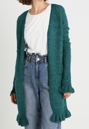 Vrouw draagt een teal gebreid vest met ruches aan de manchetten en zoom, over een wit shirt en high-waisted blauwe jeans met een geplooide tailleband.
