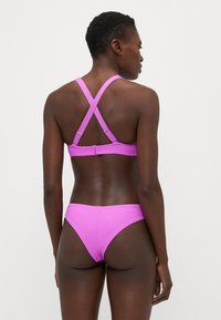 Rožinis bikini komplektas, pasižymintis tekstūruotu dizainu, reguliuojamomis kryžminėmis petnešomis ir sagtimi nugaroje. Paprasto, minimalistinio kirpimo bikinio dugnas.