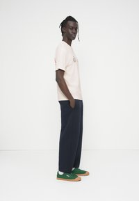 Μπεζ βαμβακερό μπλουζάκι με κόκκινο κείμενο, φαρδιά ναυτικά μπλε παντελόνια και πράσινα αθλητικά παπούτσια με καφέ σόλες. Το μοντέλο στέκεται πλαγίως, με τα χέρια στις τσέπες.