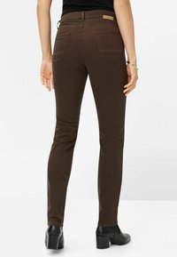 Pantalon marron slim, confectionné en tissu extensible, avec deux poches arrière et une étiquette à languette sur la ceinture. Texture lisse et design élégant.