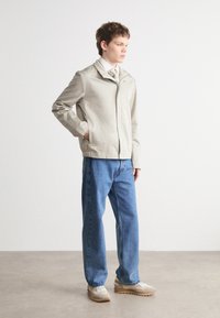 Lys grå jakke med stående krage, frontglidelås og to sidelommer, kombinert med løse blå denim jeans og beige treningssko.