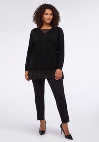 Czarna bluzka z długim rękawem i dekoltem w serek, z przezroczystym wstawką w groszki, nałożona na czarną plisowaną spódnicę, w połączeniu z dopasowanymi czarnymi spodniami i szpilkami.