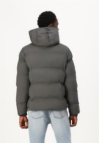 Grå pufferjakke med hætte, der har et quiltet design, tykkem nedlukning og elastiske manchetter. Bæres over lyseblå jeans.