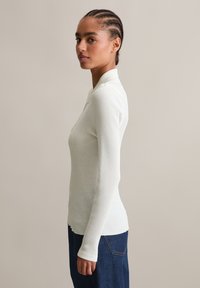 Jonge vrouw met gevlochten haar die zijwaarts staat en een strakke witte geribbelde longsleeve en een paar blauwe jeans draagt, tegen een eenvoudige achtergrond.