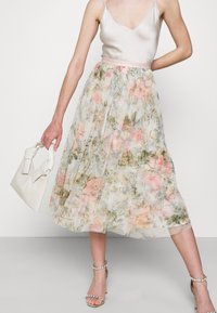 Bloemenplissérok van doorschijnende stof met roze en groene tinten, gecombineerd met een satijnen tanktop en een kleine off-white handtas.