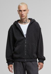 Crna zip-up hoodie s teksturiranim uzorkom, s velikom kapuljačom, prednjim džepovima i rebrastim rukavima. Nosi se preko bijele majice.
