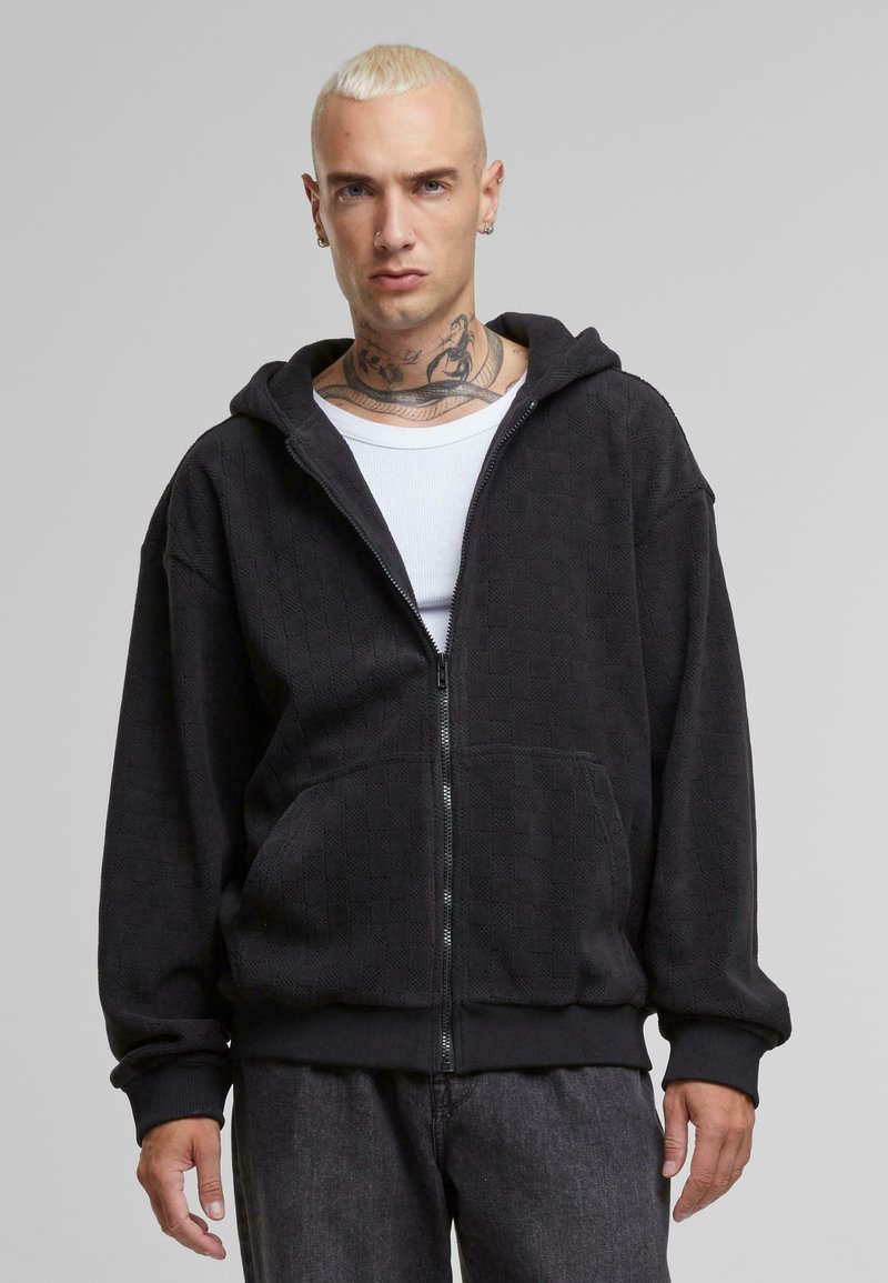 Crna zip-up hoodie s teksturiranim uzorkom, s velikom kapuljačom, prednjim džepovima i rebrastim rukavima. Nosi se preko bijele majice.
