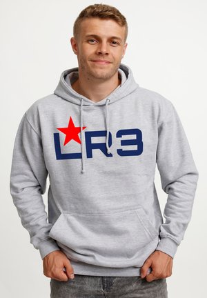 Young man wearing a light gray hoodie with dark blue "LR3" text and a large red star on the chest, standing against a plain background.