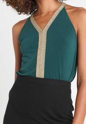 Vrouw draagt een mouwloos groen topje met een metalen gouden V-vormige rand, gecombineerd met een zwarte rok met hoge taille.