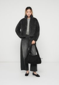 Kvinde iført en mørkegrå fleecejakke, brede, falmede sorte jeans, sorte flade sko, som holder en sort rynket håndtaske mod en simpel lys baggrund.