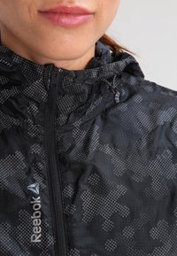 Czarna kurtka Reebok z teksturowanym wzorem, zamkiem błyskawicznym, regulowanym kapturem i odblaskowymi detalami. Tkanina wydaje się lekka i oddychająca.