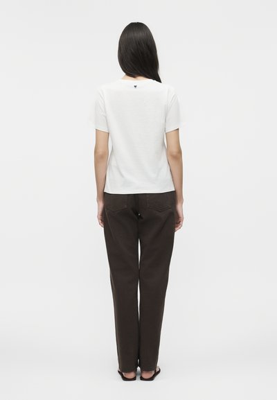 Γυναίκα με μακριά σκούρα μαλλιά στέκεται γυρισμένη προς τα πίσω, φοράει απλό λευκό κοντομάνικο πουκάμισο, καφέ σκούρο παντελόνι και μαύρες σαγιονάρες.