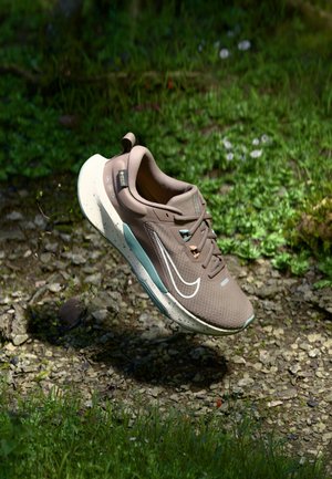 Chaussure de course Nike marron clair avec semelle blanche et accents bleu sarcelle, suspendue au-dessus de gravier et d'herbe en plein air sous la lumière du soleil.