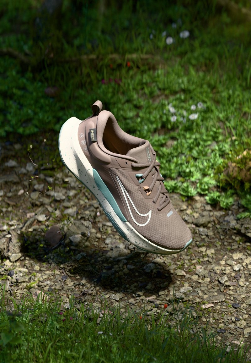 Chaussure de course Nike marron clair avec semelle blanche et accents bleu sarcelle, suspendue au-dessus de gravier et d'herbe en plein air sous la lumière du soleil.