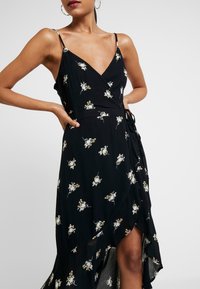 Vestido negro ajustado con tirantes finos, presenta un patrón floral en blanco y amarillo, con un dobladillo asimétrico y un detalle de lazo lateral para el cierre.
