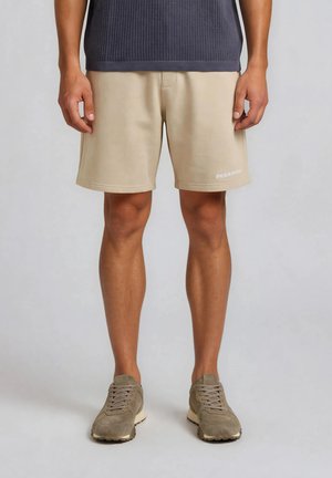 Mand iført beige shorts og taupe sneakers, stående med armene afslappet ned langs siderne mod en ensartet lys baggrund.