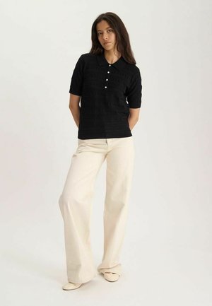 Jeune femme debout portant un polo noir à manches courtes et un pantalon large crème sur un fond clair uni.