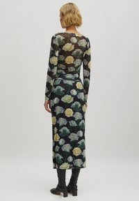 Langärmliges, transparentes Kleid mit schwarzem Grund, das ein florales Muster aus gelben und grünen Rosen zeigt. Kombiniert mit kniefreien schwarzen Stiefeln.