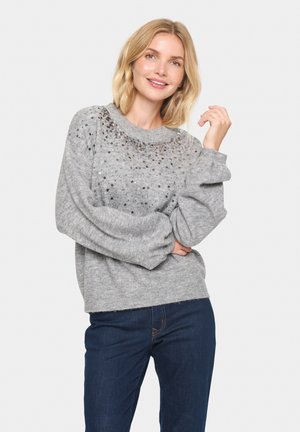 Pull en tricot gris avec des manches ballon, présentant un design à paillettes sur la poitrine. Associé à un jean en denim bleu foncé.