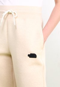 Beige sporta bikses ar auklu jostasvietā, sānu kabatām un melnu gumijas logotipa uzšuvumu kreisajā augšstilbā. Gluda, mīksta tekstūra.