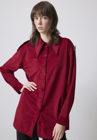Giovane donna con capelli castani fino alle spalle, che indossa una lunga camicia di velluto a coste rossa con colletto ampio, in piedi davanti a uno sfondo semplice.