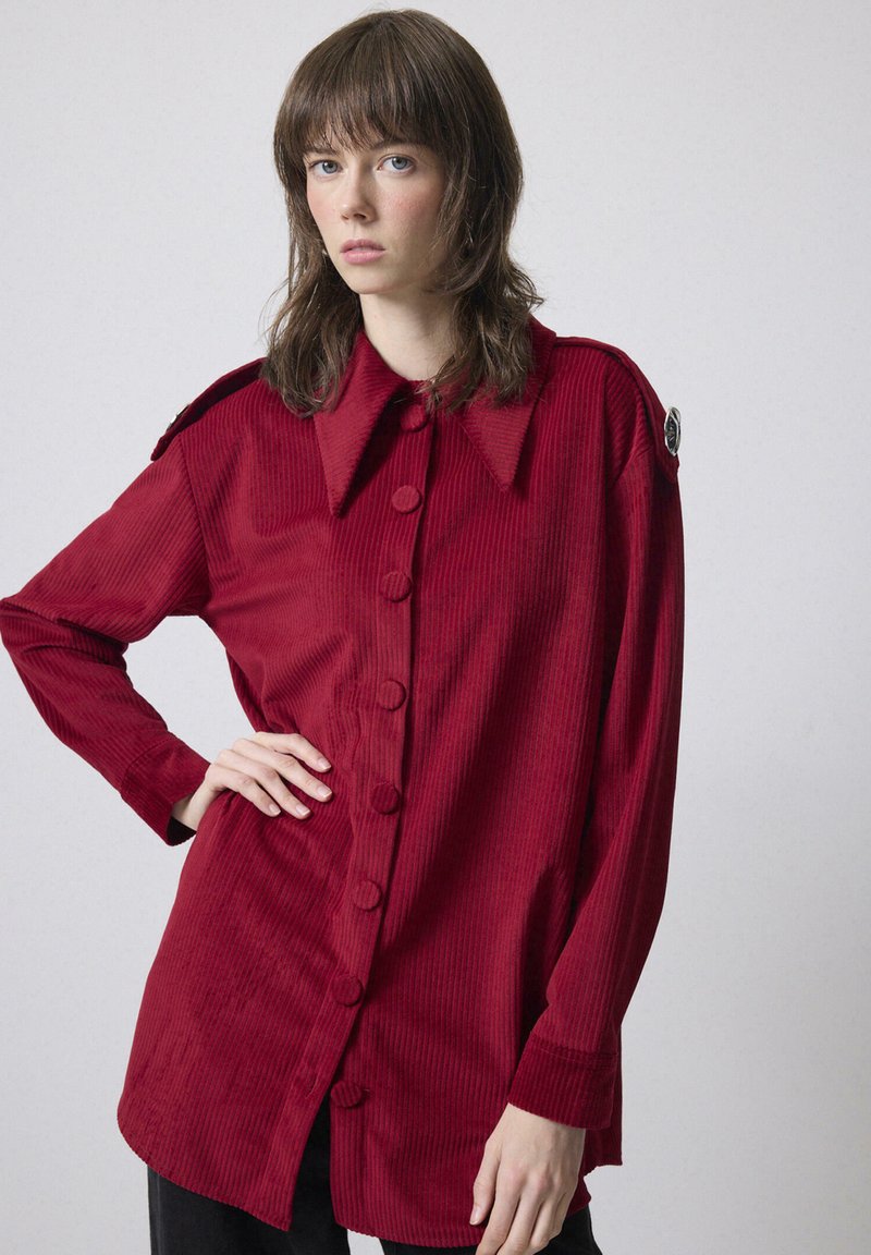 Giovane donna con capelli castani fino alle spalle, che indossa una lunga camicia di velluto a coste rossa con colletto ampio, in piedi davanti a uno sfondo semplice.