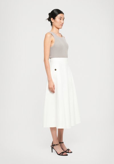 Woman standing in grey sleeveless top and white midi skirt with pocket detail, wearing black strappy heels, against plain white background.