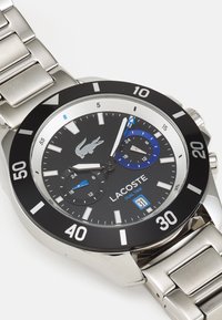 Reloj de acero inoxidable con esfera negra, acentos plateados, doble pantalla de hora y elementos azules. Cuenta con un bisel audaz y pulsera de eslabones.