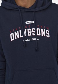 Sudadera azul marino con cordones, con el texto blanco y burdeos "ONLY&SONS", diseño de mezclilla vintage y un bolsillo frontal.