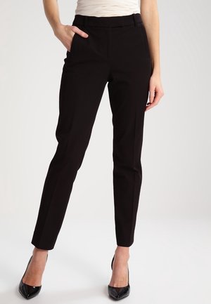 Nainen pukeutuneena istuviin mustiin housuihin ja mustiin korkokenkiin, toinen käsi taskussa, seisoo yksiväristä taustaa vasten.