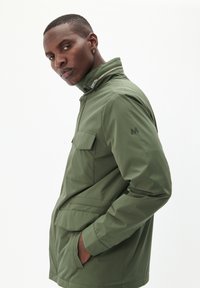 Olivgrüne Jacke mit hohem Kragen, Fronttaschen und einem Reißverschlussdetail. Besitzt ein dezentes Logo auf dem Ärmel. Glattes, leichtes Material.