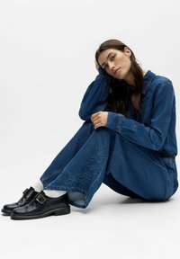 Femme aux longs cheveux bruns assise les jambes croisées, portant une tenue en denim bleu avec des broderies florales, des chaussettes blanches et des chaussures noires à boucles.