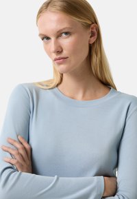 Blondynka z niebieskimi oczami, ubrana w jasnoniebieską koszulę z długim rękawem, z założonymi rękami, patrząc lekko na bok na tle jednolitego białego tła.