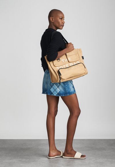 Tan canvas tote bag with white text, front pockets, and a zippered compartment. Paired with a black sweater and denim skirt. Simple design.