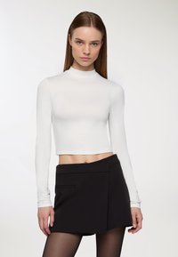Biała dopasowana bluzka z długim rękawem i wysokim kołnierzem, połączona z czarną spódnicą o kroju kopertowym z boczną kieszenią. Gładka faktura, krótka konstrukcja.