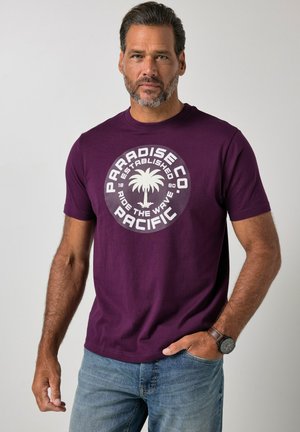 Mężczyzna o krótkich włosach i brodzie, ubrany w fioletowy t-shirt z białym logo drzewa palmowego i napisem "Paradise Co. Pacific" oraz niebieskie dżinsy.