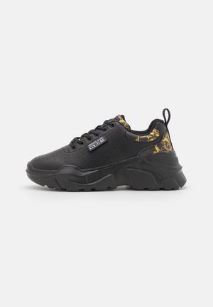 Zapatilla gruesa negra con detalles de patrón barroco dorado, laterales perforados, cordones delanteros y logo de Versace Jeans Couture en el lateral.