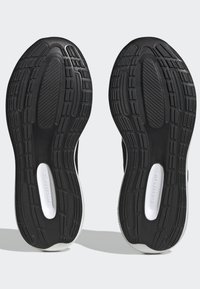 Suelas de zapatos de goma negra con surcos profundos, que incluyen patrones texturizados y zonas de amortiguación. Presenta un acento blanco de CLOUDFOAM a lo largo del costado.