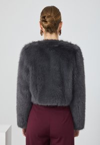 Mujer con un moño de pelo castaño lleva una chaqueta de piel gris oscuro y pantalones color burdeos, de espaldas contra un fondo claro y liso.
