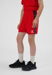 Rode sportieve korte broek met een zwarte chevronstreek langs de zijkant, een elastische tailleband en een wit logodetail. Zwarte schoenen en lichte sokken maken de outfit compleet.