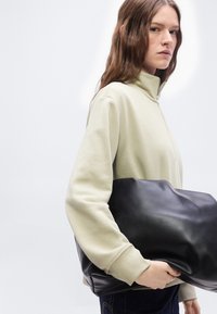 Γυναίκα με μακριά καστανά μαλλιά που φοράει ένα ανοιχτό μπεζ πουλόβερ με φερμουάρ, κρατώντας μια μεγάλη μαύρη δερμάτινη τσάντα φάκελο πάνω σε λευκό φόντο.