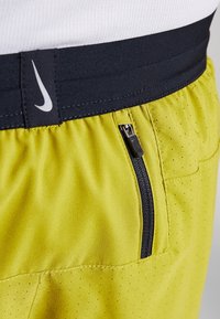 Nærbillede af gule atletikshorts med sort elastisk taljebånd med hvid Nike-logo og en lodret sort lynlåslomme på siden.