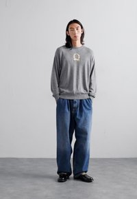 Grijze sweatshirt met een geborduurd ontwerp, gecombineerd met losse blauwe denim jeans. De outfit wordt aangevuld met zwarte schoenen en heeft een gestructureerde uitstraling.
