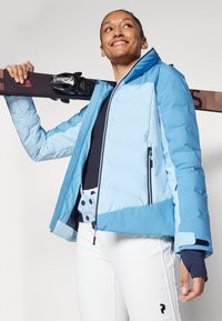 Blue ski jacket with a zippered front, contrasting dark accents, and a soft texture. Paired with white ski pants featuring button details.