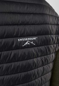 Gros plan d'une veste noire matelassée avec des coutures horizontales et le logo Swedemount au-dessus d'un motif de montagne blanche dans le dos.