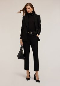 Zwart getailleerd blazer met een slanke silhouette, gecombineerd met strakke zwarte broek en een doorzichtige zwarte turtleneck; accessoires omvatten een zwarte handtas.