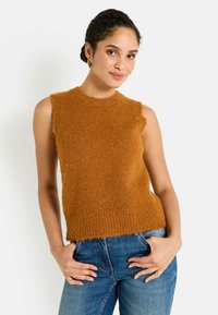 Femme avec des cheveux tressés portant des boucles d'oreilles en or, un pull sans manches en peluche marron et un jean bleu, souriant avec les mains dans les poches.