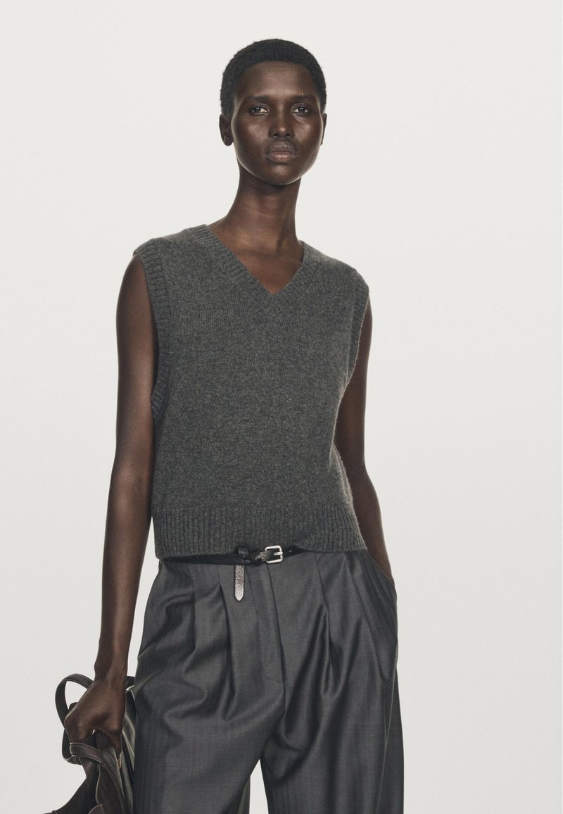 Sleeveless gray knit vest with a V-neck, paired with high-waisted, pleated dark gray trousers and a black belt, holding a brown bag.
